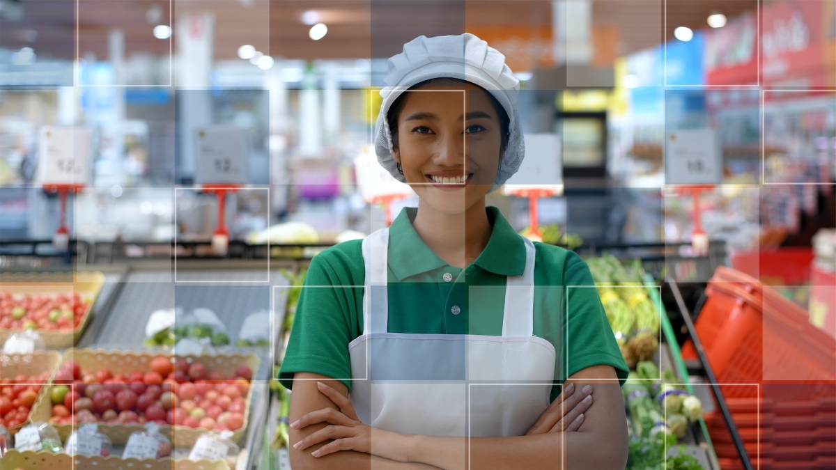 A female grocery store clerk smiles while standing in front of some fruit and vegetable stands